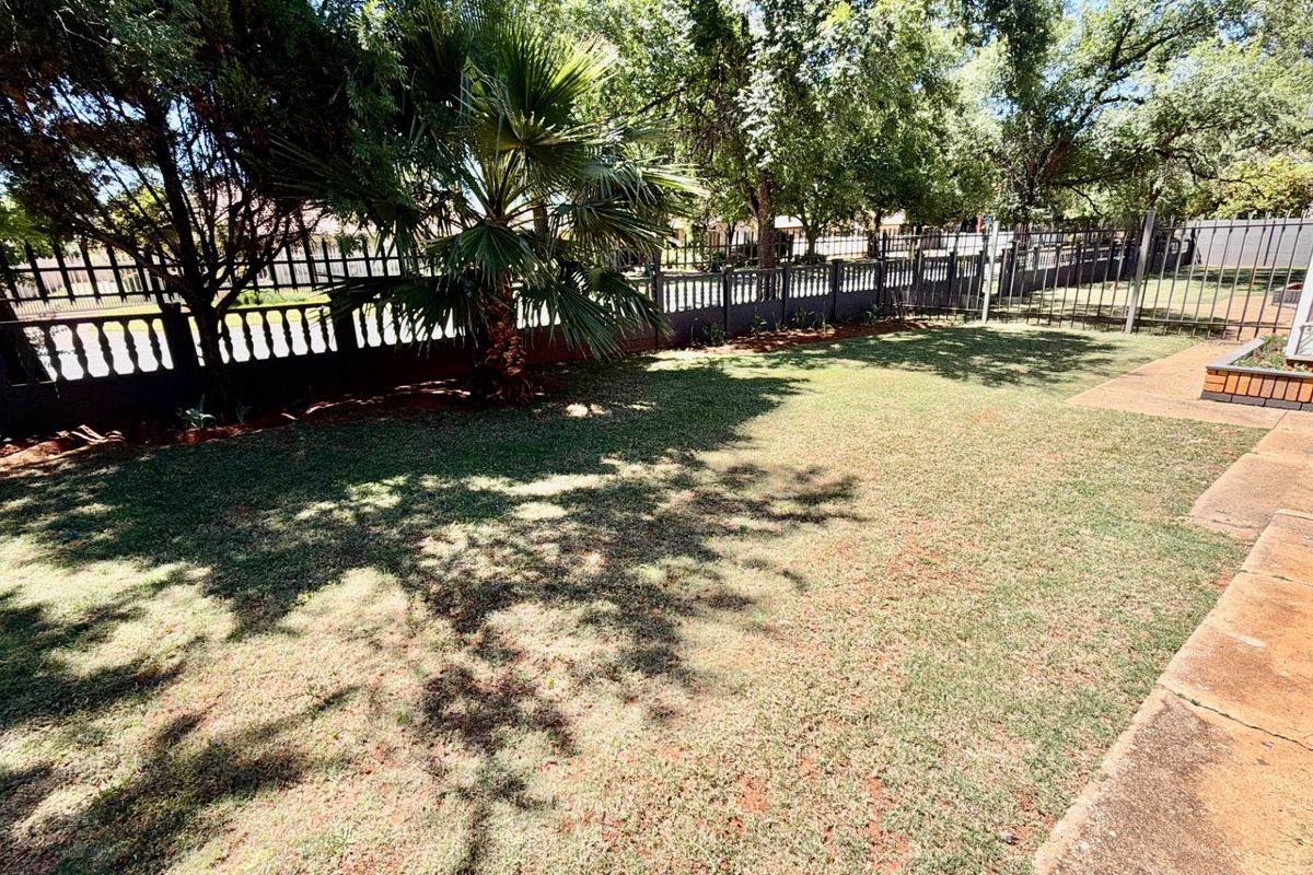 A well-maintained rear garden featuring a lawn, mature trees, and metal perimeter fencing, adjacent to a paved patio area.