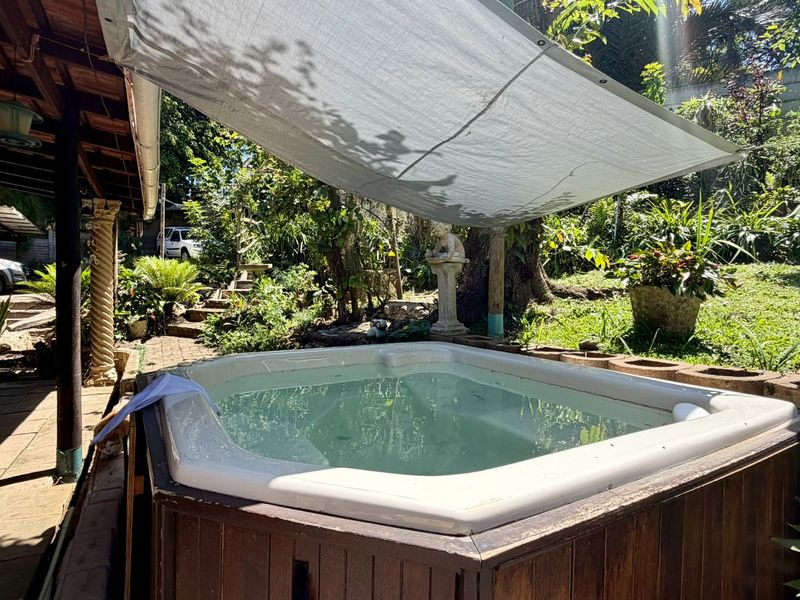 Outdoor area featuring a built-in spa with timber cladding, situated under a covered patio with a shade sail and surrounding garden.