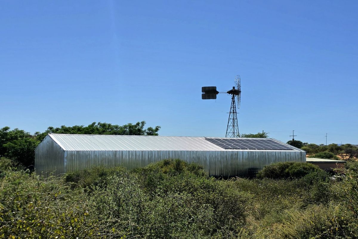A large corrugated metal farm shed featuring roof-mounted solar panels and an adjacent traditional wind pump, set within a rural landscape.