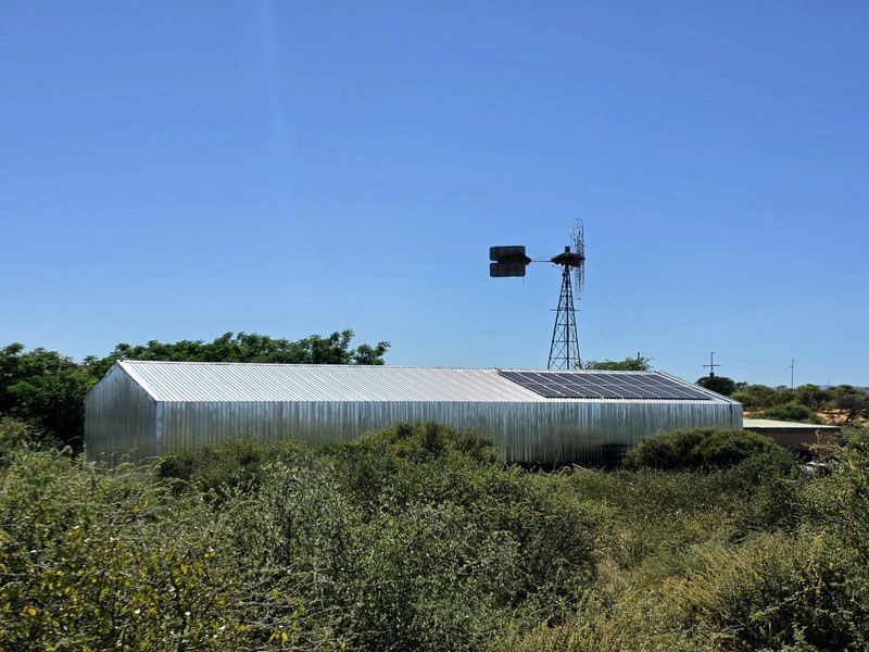 A large corrugated metal farm shed featuring roof-mounted solar panels and an adjacent traditional wind pump, set within a rural landscape.