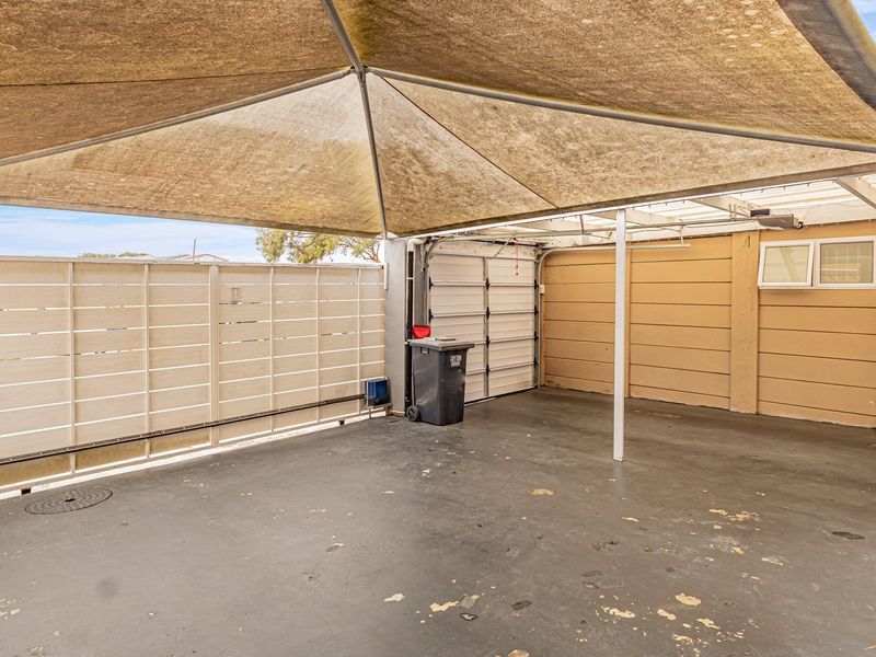 A covered outdoor parking area or garage space featuring a large shade sail, concrete flooring, and secure metal perimeter fencing.