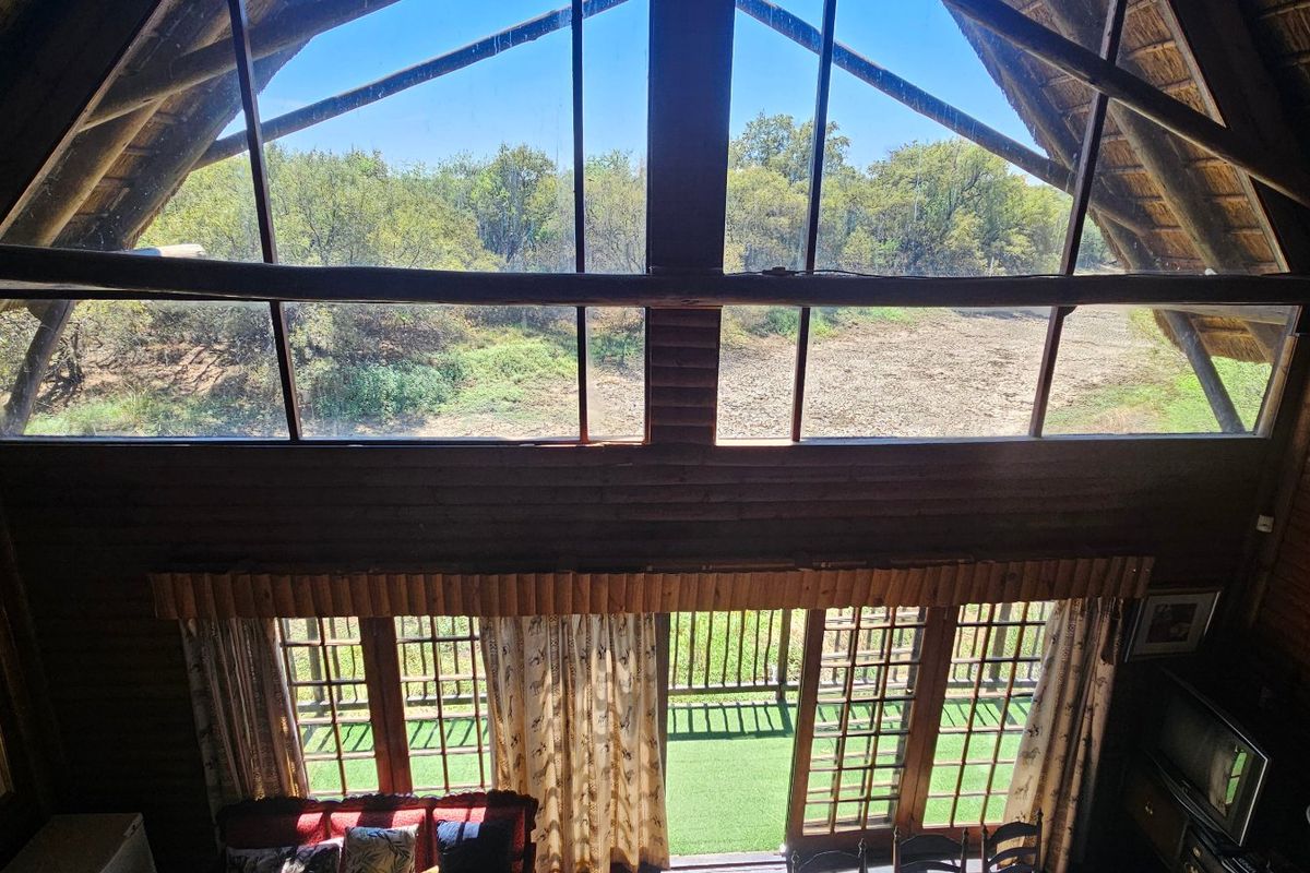 Spacious double-volume living area featuring rustic log walls, exposed timber beams, a traditional thatch roof, and expansive windows offering scenic landscape views.