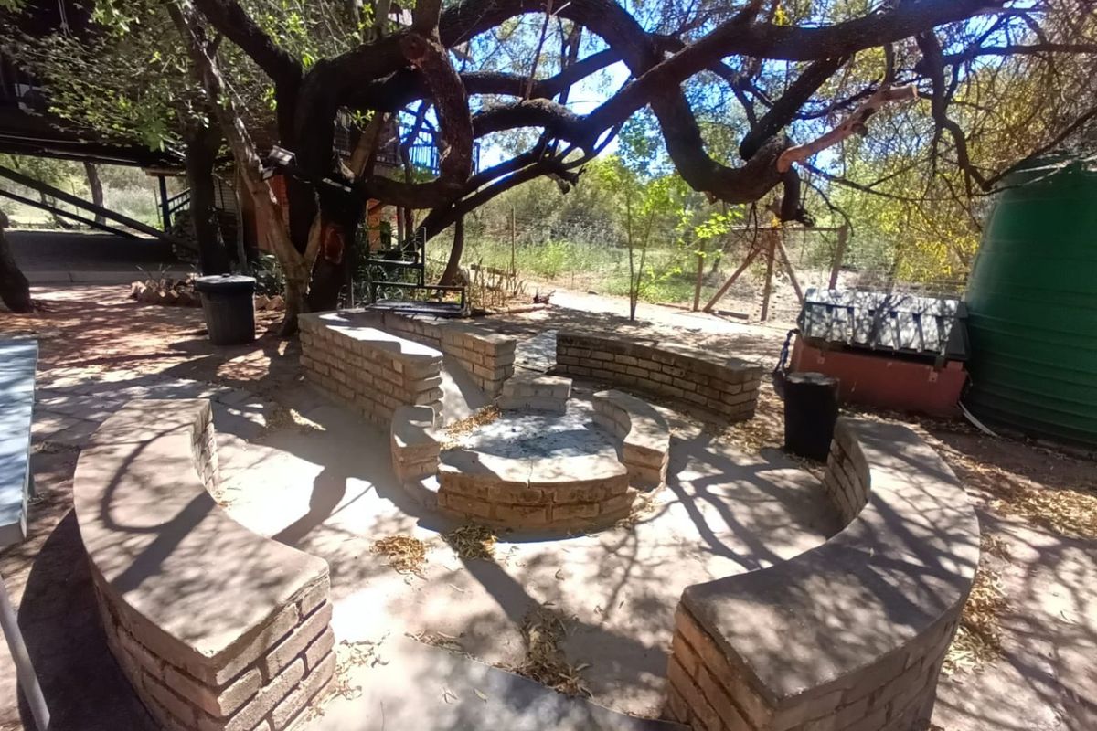 Outdoor entertainment area featuring a built-in brick fire pit with curved masonry seating, situated under mature trees in a natural garden setting.