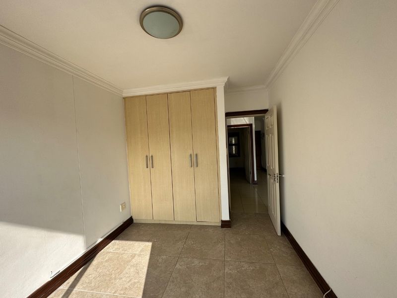 A clean, empty bedroom featuring neutral tiled flooring, built-in wooden cupboards, and classic ceiling coving, offering a versatile space ready for personalization.