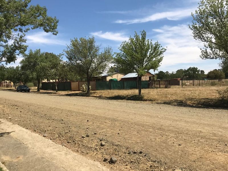 A street-level view of a residential plot featuring a gravel road, concrete sidewalk, and perimeter fencing surrounding the vacant land.