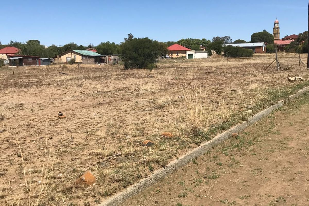 A vacant residential plot featuring dry grass, wire fencing, and a concrete curb, offering a clear view of the surrounding neighborhood.