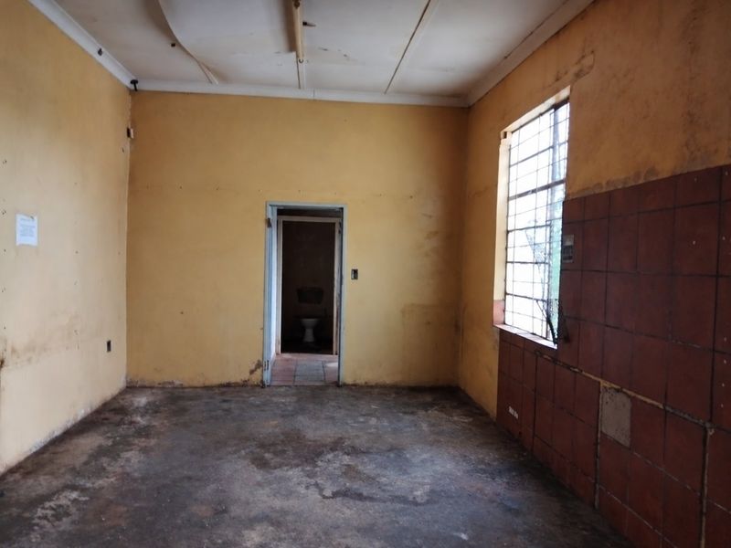 An empty, dilapidated utility space featuring security bars on the window, partial wall tiling, and significant wear to the floor and wall finishes.