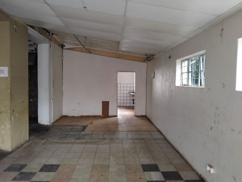 An empty, open-plan commercial space featuring tiled flooring, a suspended ceiling, and security-barred windows, requiring significant renovation and cosmetic updates.