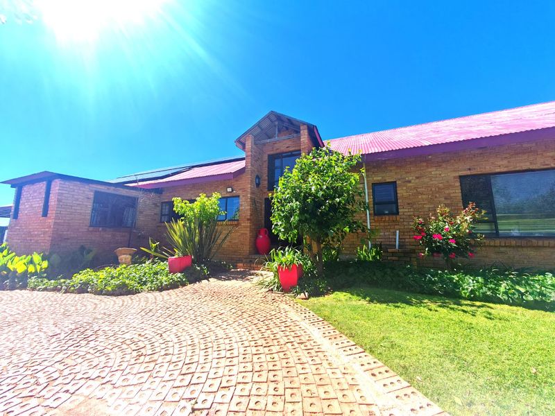 A well-maintained face brick farmhouse featuring a distinctive red corrugated metal roof, solar panels, and a circular paved driveway entrance.