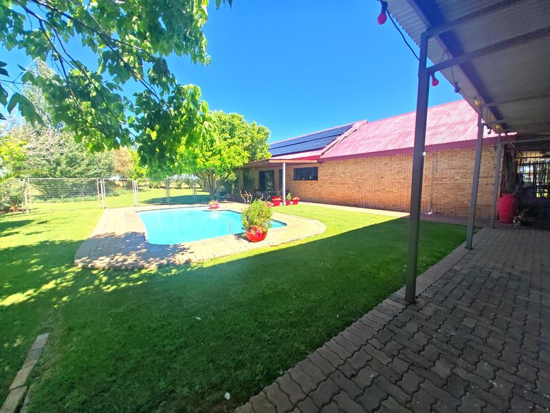 A spacious outdoor area featuring a swimming pool, manicured lawn, and a covered patio attached to a brick farmhouse with solar panels.