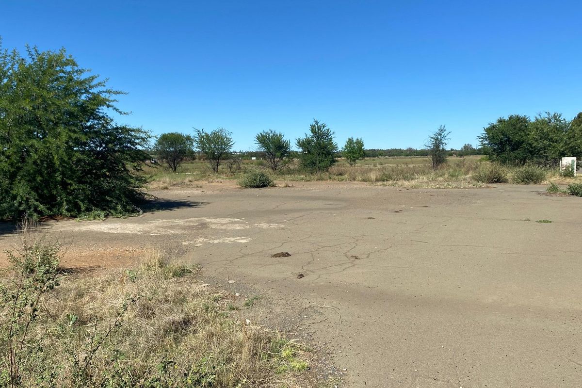 A view of a vacant residential plot featuring an existing asphalt driveway and surrounding natural vegetation under a clear blue sky.