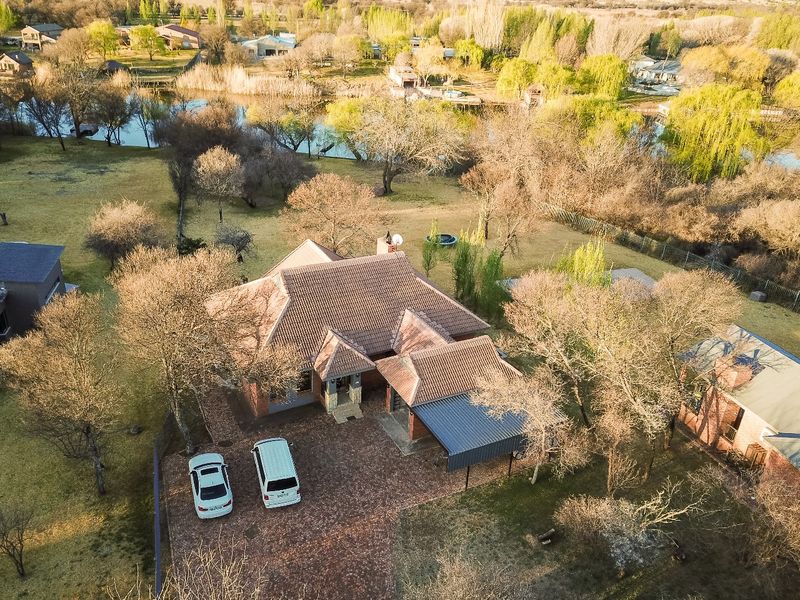 Aerial view of a residential property featuring a tiled roof, brick driveway, carport, and a spacious garden setting near a water feature.