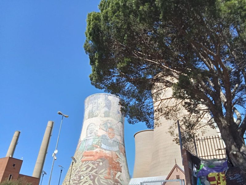 Exterior view of industrial cooling towers featuring a large mural, set within a fenced commercial site.