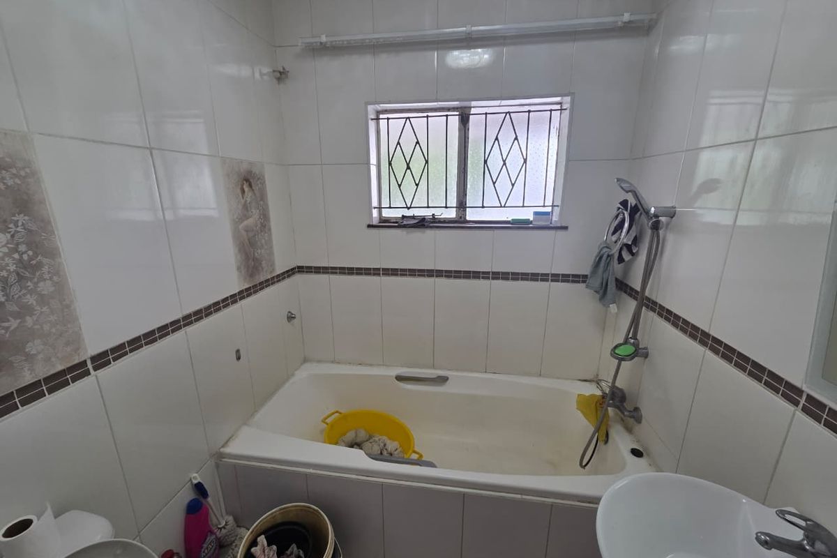 A dated bathroom featuring a built-in bathtub, wall-mounted shower mixer, and pedestal basin, finished with ceramic wall tiles and security bars on the window.
