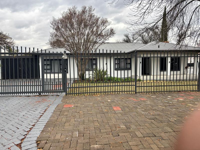Front exterior view of a single-story house featuring a paved driveway, secure metal fencing, and a corrugated metal roof.