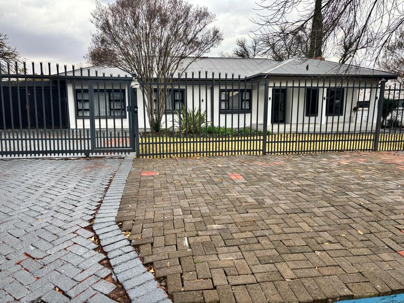 Front exterior view of a single-story house featuring a paved driveway, secure metal fencing, and a modern corrugated metal roof.