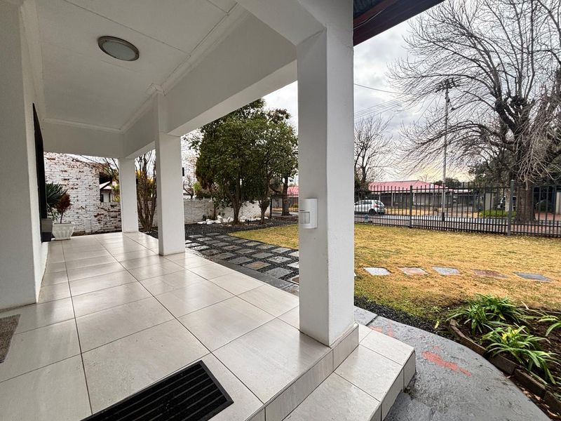 A covered patio area featuring tiled flooring and support pillars, overlooking a front garden with metal perimeter fencing.