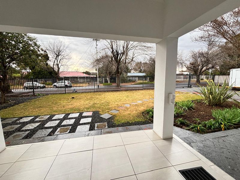 A covered patio area featuring tiled flooring and a concrete pillar, overlooking a landscaped front garden with paved stepping stones and secure perimeter fencing.