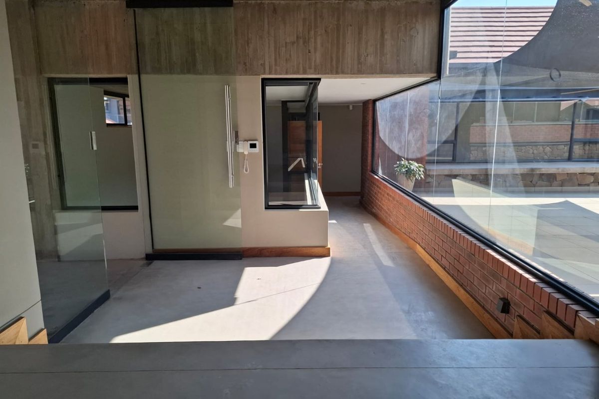 Modern open-plan living area featuring exposed concrete ceilings, brick accents, and expansive floor-to-ceiling windows overlooking an outdoor patio space.