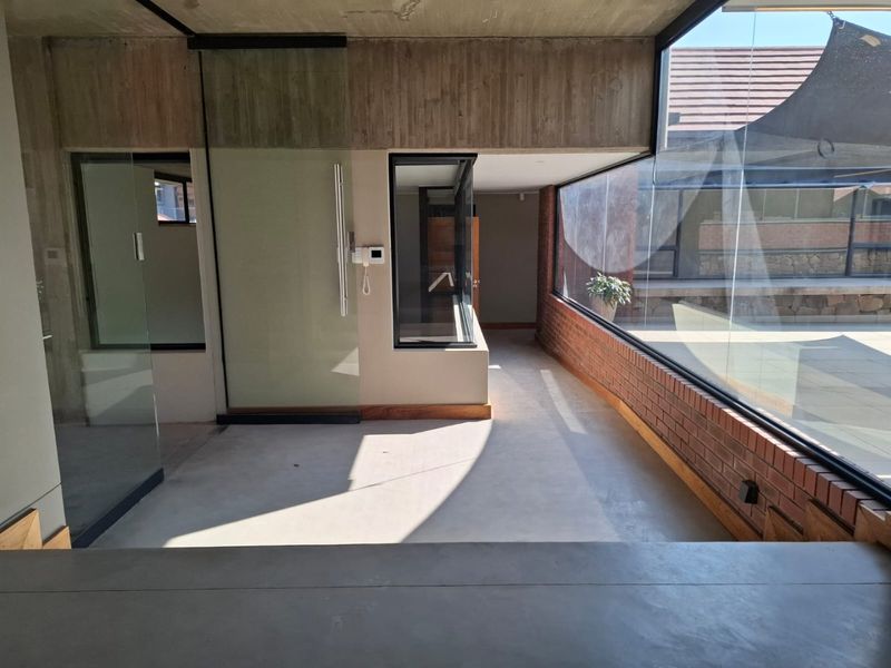 Modern open-plan living area featuring exposed concrete ceilings, brick accents, and expansive floor-to-ceiling windows overlooking an outdoor patio space.