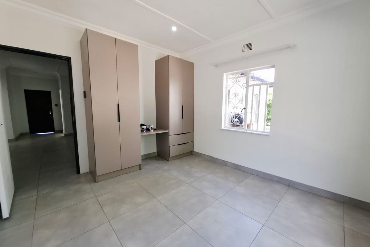 A clean, empty bedroom featuring neutral tiled flooring, modern built-in cupboards with an integrated desk, and a window with security bars.