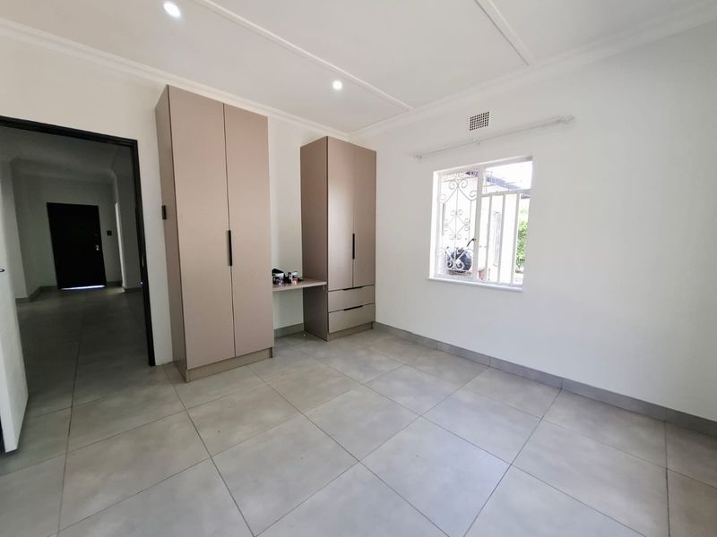 A clean, empty bedroom featuring neutral tiled flooring, modern built-in cupboards with an integrated desk, and a window with security bars.