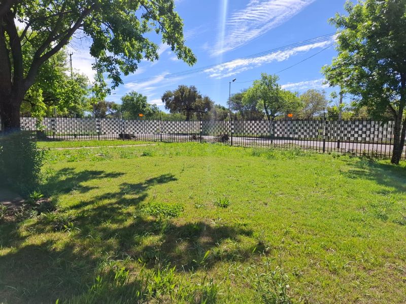 A spacious, open garden area featuring a well-maintained lawn, mature trees, and secure metal perimeter fencing.