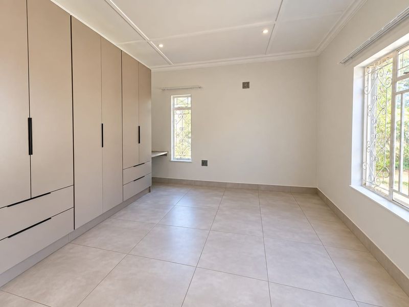 A bright, empty bedroom featuring neutral tiled flooring, extensive built-in cupboards with a integrated desk, and security bars on the windows.