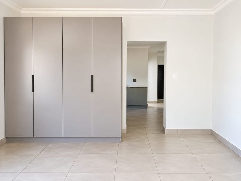 A clean, modern bedroom featuring neutral tiled flooring, crisp white walls, and a large, contemporary built-in wardrobe with matte grey finish.
