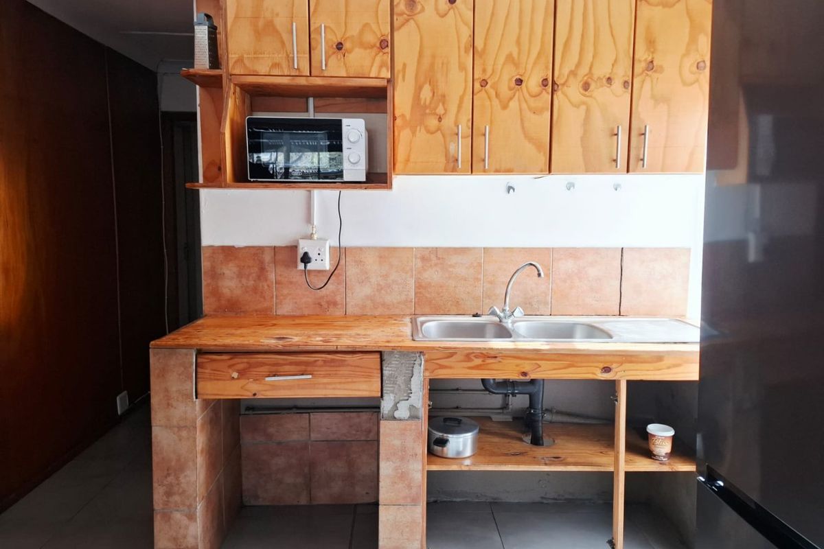 A functional kitchen featuring wooden cabinetry, a double bowl sink, and tiled splashbacks, currently showing signs of wear and unfinished construction details.
