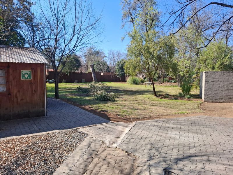 A spacious garden area featuring a paved driveway, a wooden storage shed, and mature trees, enclosed by a brick boundary wall.