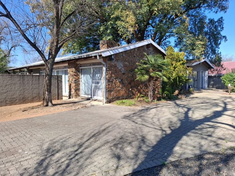 Front exterior view of a stone-clad house featuring a paved driveway and security gates, set within a mature garden environment.