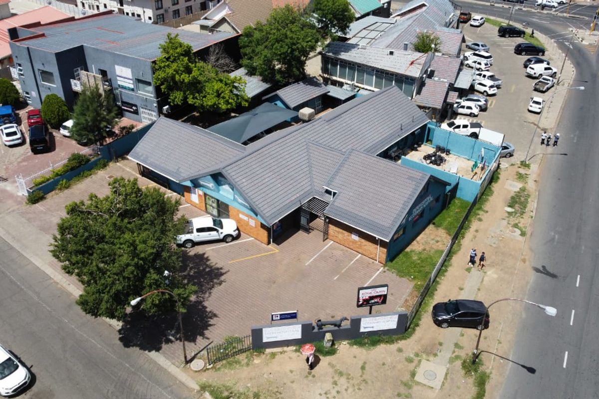 Aerial view of a commercial property featuring a tiled roof building, paved parking area, and perimeter security fencing situated near a main road.