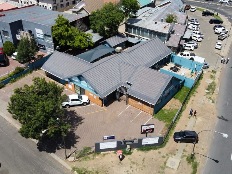 Aerial view of a commercial property featuring a tiled roof building, paved parking area, and perimeter security fencing situated near a main road.