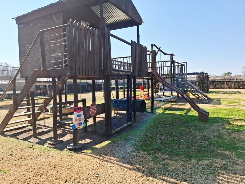 A large wooden jungle gym structure situated in a spacious garden area, featuring a slide, climbing net, and elevated play platform.