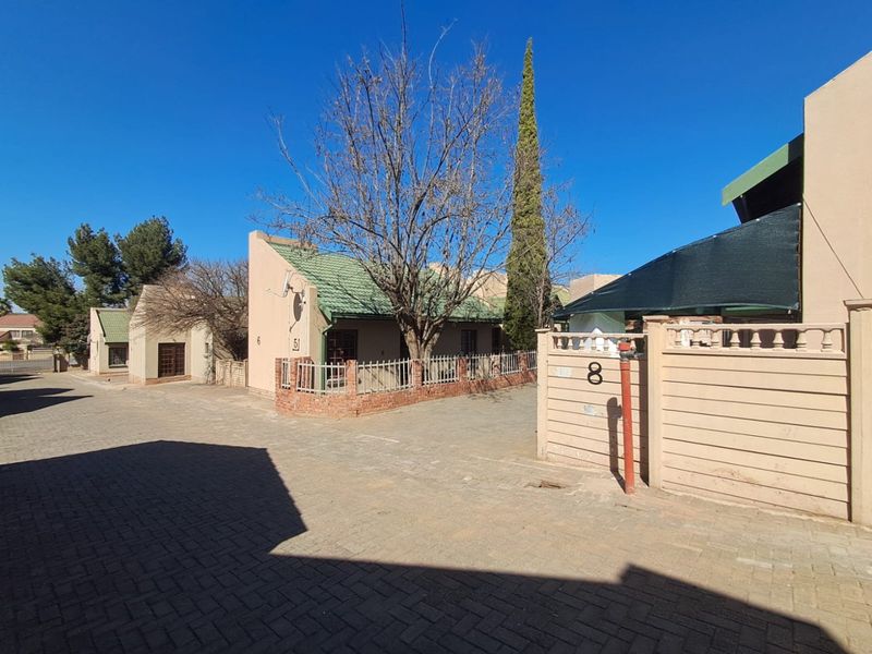 A street-level view of a townhouse complex featuring paved driveways, brick boundary walls, and tiled roof structures under a clear sky.
