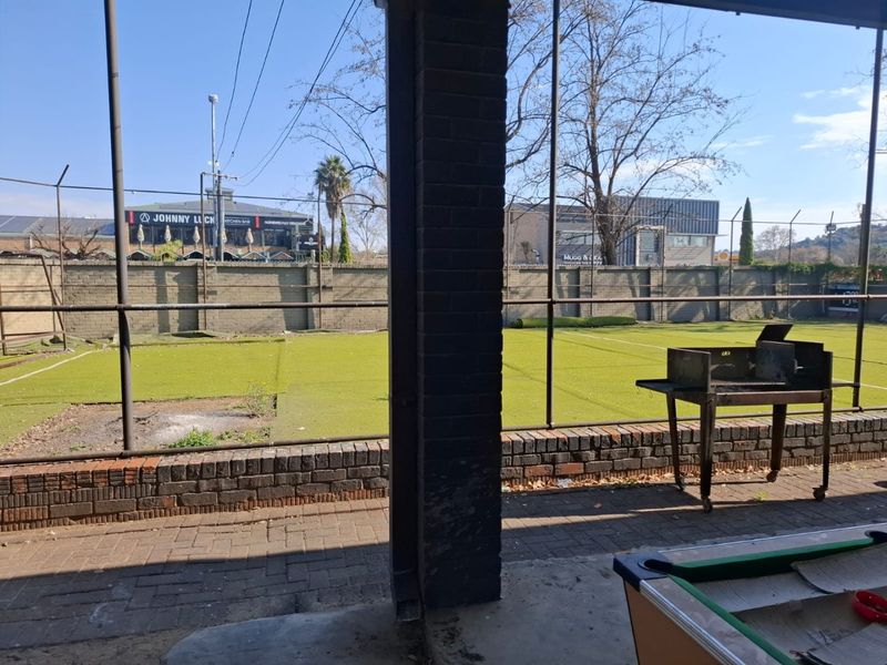 A covered patio area featuring brick paving and a brick pillar, overlooking a large outdoor space with artificial turf and perimeter fencing.