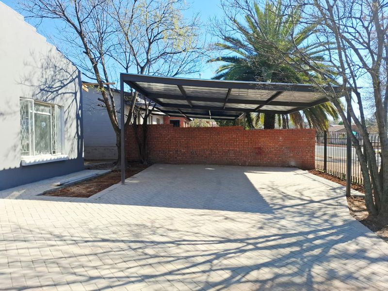 A well-maintained front exterior featuring a paved driveway and a modern steel-framed carport, enclosed by a brick boundary wall and metal fencing.