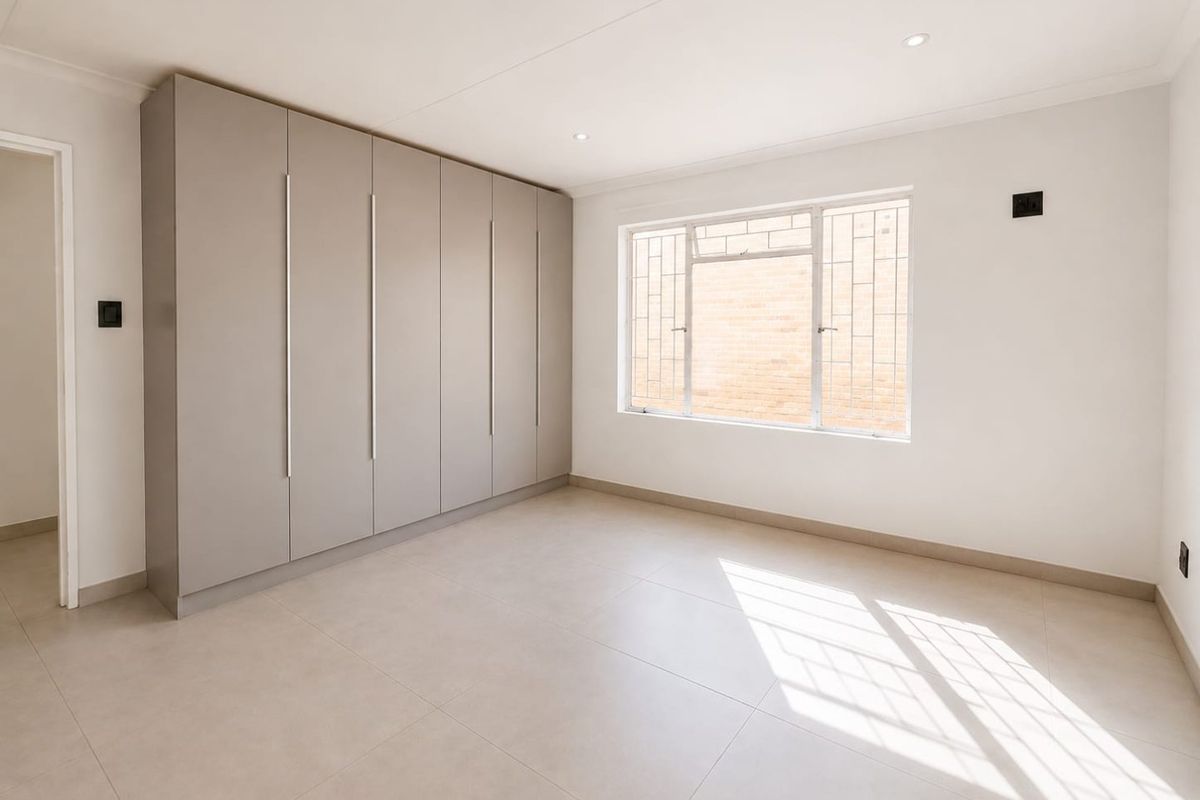 A bright, neutral bedroom featuring modern tiled flooring, recessed downlighting, and extensive built-in cupboard storage with a window fitted with security bars.