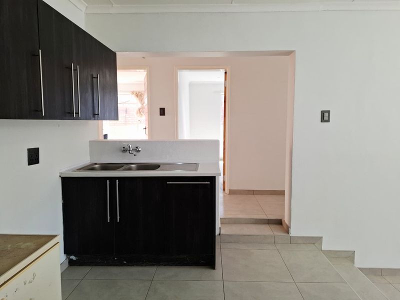 A compact kitchen area featuring dark built-in cabinetry, a double sink with mixer tap, and neutral floor tiles, leading into an adjacent living space.