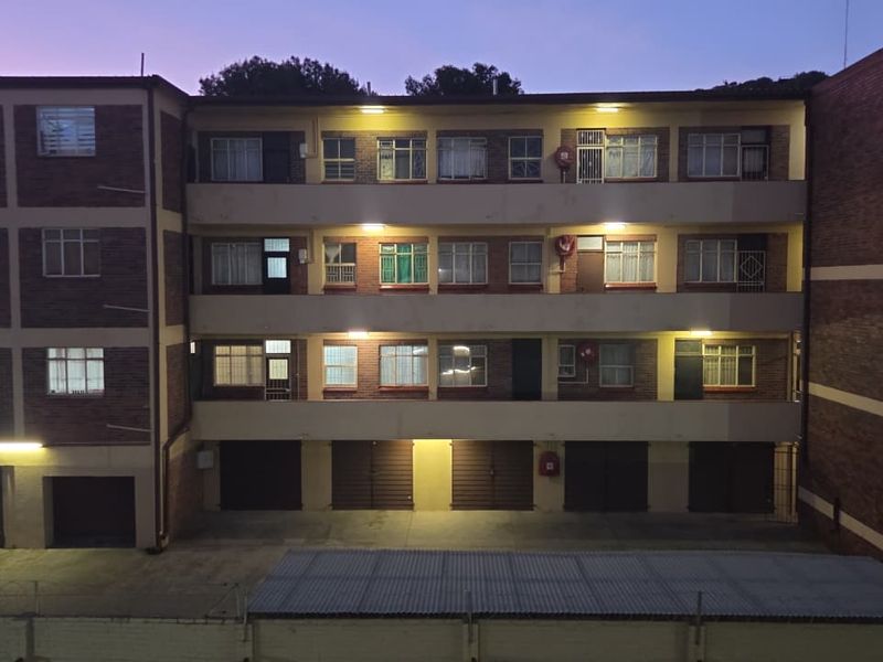 A multi-story apartment building exterior featuring brick facade, balcony access walkways, and ground-level garage units at dusk.