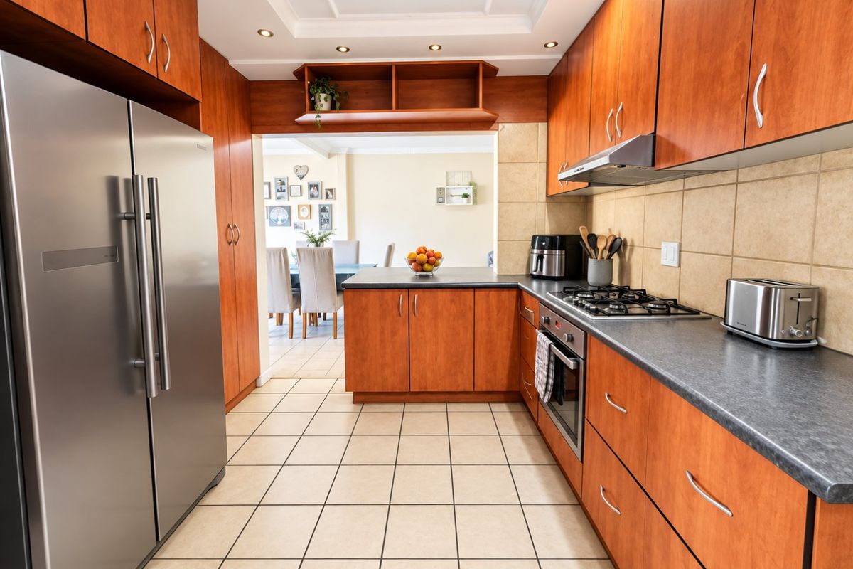 A functional kitchen featuring warm-toned wooden cabinetry, dark laminate countertops, a gas hob, and tiled flooring, with an open-plan flow to the dining area.