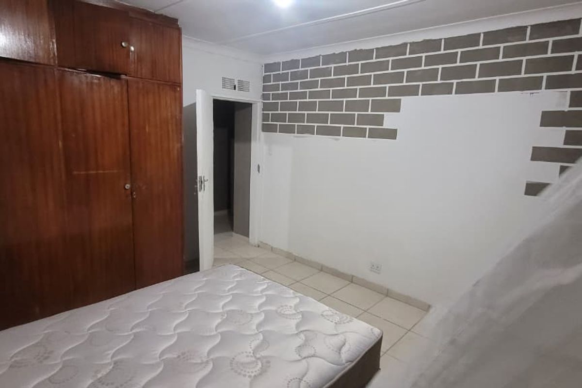 Bedroom featuring built-in wooden cupboards, tiled flooring, and an unfinished exposed brick feature wall requiring cosmetic attention.