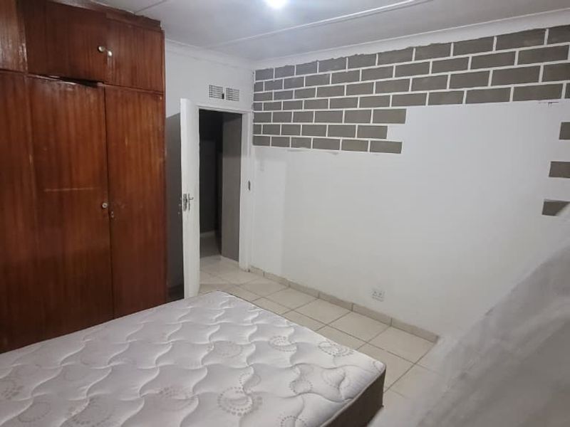 Bedroom featuring built-in wooden cupboards, tiled flooring, and an unfinished exposed brick feature wall requiring cosmetic attention.
