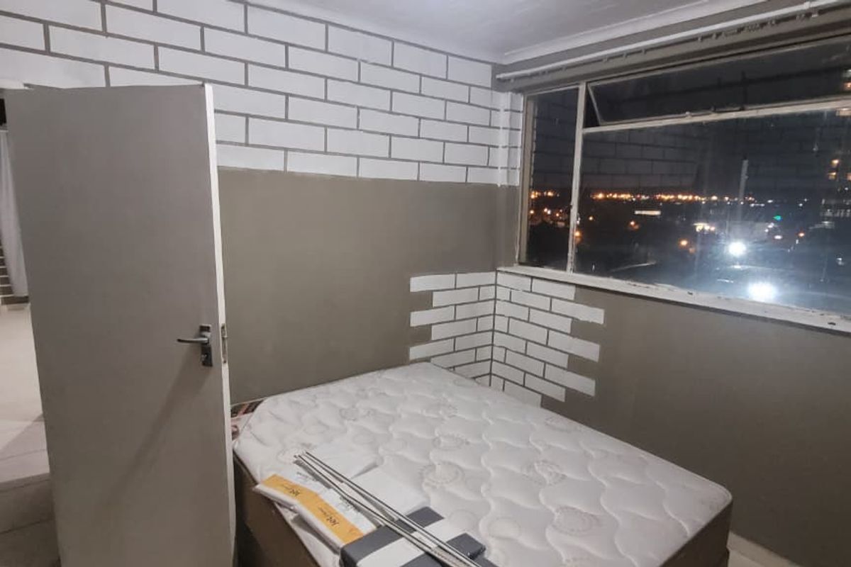 A bedroom featuring large windows with city views, tiled flooring, and a partially painted brick-effect wall finish.