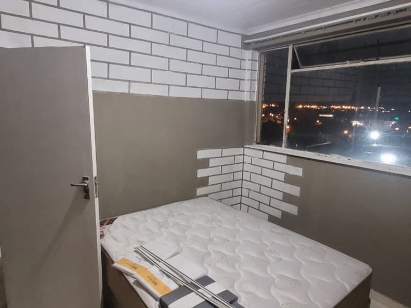 A bedroom featuring large windows with city views, tiled flooring, and a partially painted brick-effect wall finish.