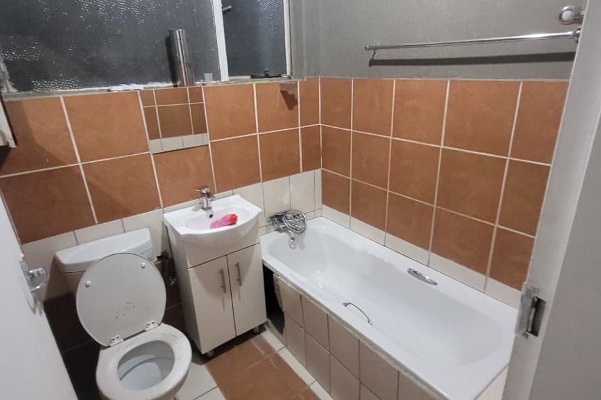 A compact bathroom featuring a bathtub, toilet, and vanity basin with brown wall tiling and tiled flooring, requiring some cosmetic updates.