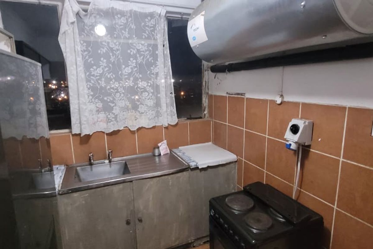A compact kitchen featuring a stainless steel sink, tiled splashback, and a wall-mounted electric geyser, requiring cosmetic updates to improve functionality and aesthetic appeal.