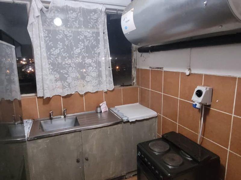 A compact kitchen featuring a stainless steel sink, tiled splashback, and a wall-mounted electric geyser, requiring cosmetic updates to improve functionality and aesthetic appeal.