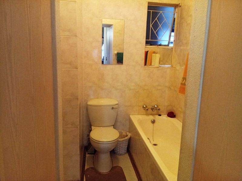 A compact, dated bathroom featuring a built-in bathtub, toilet, and full-height wall tiling, requiring modernization to improve its aesthetic appeal.